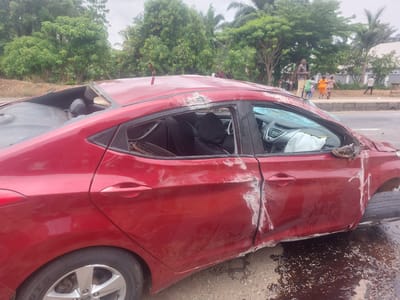 The image depicts a severely damaged red sedan on the side of a road in Akwanga, Nigeria. The vehicle's roof is crushed, windows are shattered, and there is extensive damage to the side panels, particularly around the front passenger door. The front tire is completely deflated and detached from the car. A deployed airbag is visible inside the vehicle. The road is paved and appears wet, with a dark fluid pooling beneath the damaged car. In the background, there are trees and a group of people standing on the side of the road. They are observing the scene, indicating a possible accident site.
