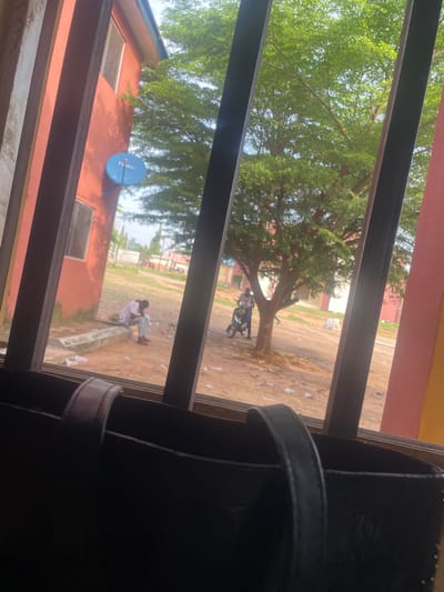 The image captures a view through a barred window, looking out onto an outdoor scene. In the foreground, a portion of a dark-colored bag with a leather strap is visible. Beyond the bars, a red building with windows is present on the left. A blue satellite dish is mounted on the side of this building.
Further into the scene, a large tree with lush green foliage dominates the middle ground. To the left of the tree, a person is seated on a concrete structure, appearing to be in a dejected pose, with their head bowed. To the right of the tree, another person is standing next to a motorcycle. The ground is sandy and somewhat littered with debris. In the background, other buildings and some distant figures can be observed, suggesting a populated area, possibly a school compound or residential area within Lokoja, Nigeria. The lighting suggests it is daytime, with the sky partially visible through the tree canopy.