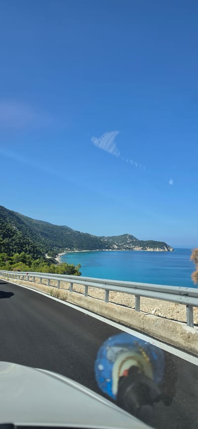This image depicts a scenic coastal road within the Municipal Unit of Lefkada, Greece, likely on the western side of the island, viewed from inside a moving vehicle on a clear, sunny day.

The upper two-thirds of the frame are dominated by a vibrant azure blue sky. In the upper right quadrant, a distinct, elongated cloud formation, resembling a stylized heart or an angular wing, is composed of wispy white cirrus, with faint, parallel linear streaks extending from it.

The horizon is an undulating line formed by densely forested green mountains meeting the sea. The midground features lush green hills and mountains sloping steeply down to the coastline. Scattered white structures are visible on the distant hillsides and a prominent headland in the mid-right, indicating some human development. The sea itself is a brilliant turquoise blue, appearing calm with lighter shades near the shore indicating shallow waters and deeper blues further out. A sandy or pebbly beach is visible at the base of the mountains in the mid-left, nestled against the coast.

In the foreground, the dark gray asphalt of a paved road sweeps from the lower left towards the right, marked by a crisp white solid line at its edge. A silver-colored, triple-beam metallic guardrail with vertical support posts runs along the right side of the road, separating it from the steep drop-off to the coast below. The road shoulder beneath the guardrail is rocky and light-colored.

The bottom left corner shows a portion of a white vehicle's interior (likely the dashboard or hood). A highly blurred and distorted reflection of a blue object, possibly a helmet or a patterned item with white dots and other colored elements, is prominently visible on the vehicle's interior surface (likely the windshield or dashboard) in the bottom right quadrant, partially obscuring the asphalt. No people are discernible in the scene.