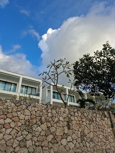 The image shows a modern white building with balconies and glass windows, seen from behind a rough stone wall. A tree with green leaves and white flowers stands in front of the building, partially obscuring it. The sky above is blue with some white clouds. This scene is consistent with the architecture and tropical vegetation found in Nusa Penida, Indonesia.