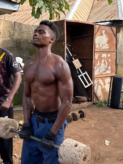 In Akwanga, Nigeria, a shirtless man with well-defined muscles is performing a strength training exercise outdoors. He is holding a barbell with what appear to be large stones as weights on either end. He is wearing blue pants and black weightlifting gloves. To his left, another person in a black and yellow shirt is partially visible. The background shows a rustic setting with a corrugated metal roof, a crumbling concrete wall, and a rusted metal cabinet containing various objects, including what looks like exercise equipment. The ground is dirt, with some leaves scattered around. The lighting suggests it is daytime.