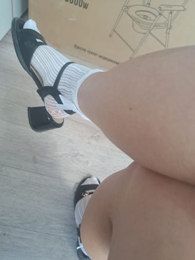 The image shows a close-up of a person's legs and feet. The person is wearing black heeled sandals with white sheer socks. The sandals have a strap across the foot with a gold buckle. The socks have a delicate lace pattern. The background is a wooden floor and a cardboard box with a drawing of a portable toilet on it. The location is Leningradskaya, Russia.