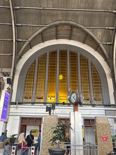The image depicts the interior of a large, arched hall, likely a train station, with a distinctive corrugated metal ceiling supported by visible metal beams and rivets. Dominating the background is a massive, arched window pane filled with amber-colored glass, divided into numerous rectangular panes by a dark grid. A large, vintage double-faced clock with Roman numerals is prominently displayed in the center of the frame, mounted on a wooden and metallic pole. Below the clock, several turnstiles are visible, with a few people interacting with them.
In the foreground and mid-ground, several individuals are present. Two adults and a child are seen near the turnstiles, one adult pointing towards a sign. Another person in a yellow hijab is walking away from the camera. To the left, a digital display board with purple and white text is mounted on a pillar. The architectural style suggests a historical building, possibly dating from the early to mid-20th century.
The overall atmosphere is one of a bustling public space, with the strong architectural elements and the presence of people indicating it is a transit hub. The amber glass of the window casts a warm, inviting glow throughout the space. The signage, particularly the red triangular sign with a fire extinguisher icon and "PEMADAM API" text, suggests this location is in Indonesia. Given the architectural style and the likely context of a major transit hub in Indonesia, this scene is likely at Gambir Station in Jakarta, Indonesia.