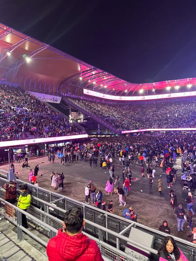 The image shows an elevated perspective of a crowd of people at Foro Sol in Mexico City, Mexico, at night. The stadium is illuminated with pink and white lights. People are walking and standing on the ground level below. Metal railings and structures are visible in the foreground.