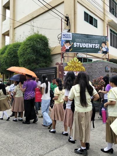 In an urban outdoor setting in Davao City, Philippines, a group of students, predominantly young girls in school uniforms, are walking on a concrete pavement. The girls are wearing beige skirts and light yellow tops with white collars and trim. Some are carrying backpacks or bags, and one is holding a bright pink water bottle.
Two adults are present: a man in a purple shirt with a reflective design, holding an orange umbrella, and another man in a green shirt. The purple-shirted man is walking alongside a student. The umbrellas are likely for protection from the sun or rain, as suggested by the overcast sky and the presence of umbrellas.
In the background, a building with a light-colored facade and windows is visible. A large banner hangs on the building, reading "NCCC Department Store WELCOME BACK STUDENTS!". Green bushy plants are situated in front of the building, adding a touch of nature to the urban landscape. The overall scene depicts students on what appears to be the first day of school or a school event.