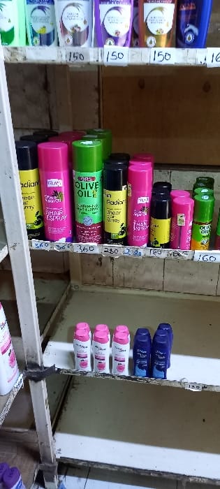 The image displays a three-tiered retail shelving unit stocked with various personal care products in what appears to be a small shop or market stall in Nairobi, Kenya.

The top shelf contains multiple plastic pouches or bottles, predominantly light green, white, purple, orange, and blue, with handwritten price tags of "150" affixed below them.

The middle shelf features an assortment of aerosol cans and smaller bottles. Visible products include "Radiant Hair Spray" in yellow and pink cans, "Ules Olive Oil Hair Spray" in a green can, and additional black and pink cans, as well as a small green bottle. Handwritten price tags beneath this shelf indicate prices of "360," "360," "360," "280," "280," "160," and "160."

The partially visible bottom shelf displays a row of five white bottles with pink caps, positioned next to a row of four dark blue bottles.

The shelving unit is constructed from a light-colored material, showing some wear and minor dust accumulation. A vertical support beam is present on the left side of the frame. The background wall is a light brown tone, and a tiled floor is visible at the bottom left. No individuals are depicted, and no actions or interactions are occurring.