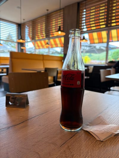 The image depicts a partially consumed bottle of Coca-Cola Zero Sugar World Cup 2026 edition, labeled with a volume of 0.33L. It is lying horizontally on a wooden table, next to a crumpled paper receipt and a metal utensil holder. The receipt contains German text. The background shows an interior space with yellow and orange striped awnings and outdoor greenery visible through windows. Based on the receipt's details referencing "Grevenmacher," the location is likely in the city of Grevenmacher, Luxembourg.