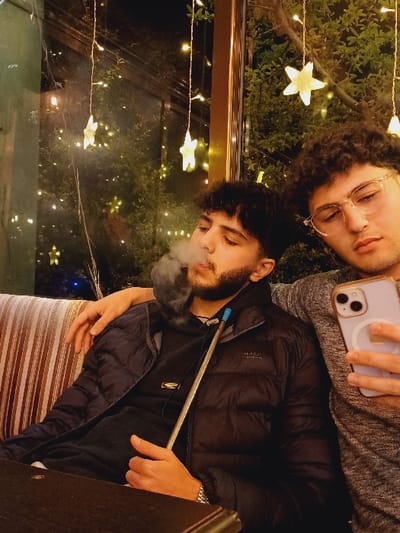 Two young men pose in a dimly lit setting adorned with star-shaped fairy lights and foliage. One, wearing glasses and a gray top, holds a phone. The other, in a black jacket, is smoking a hookah, exhaling a cloud of smoke. The image is framed within a window or glass panel, possibly at an outdoor café or lounge. The scene feels intimate and casual.