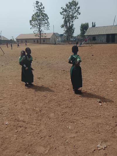 Two young girls in dark green and white school uniforms are the focus. One girl carries a smaller child also in uniform. The other stands with her back to the viewer, hands on her hips. They are on a dirt field with scattered debris. In the background are low, simple buildings with corrugated metal roofs. A few other children are visible in the distance. Trees are scattered throughout the scene under a bright sky.