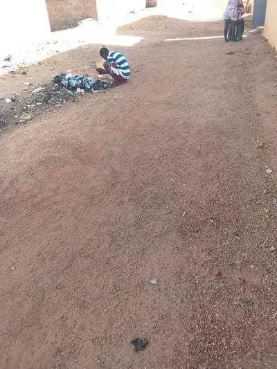 The photo shows a dirt road in Bauchi, Nigeria. In the distance, a person wearing a blue and white striped shirt and red shorts is kneeling next to a pile of trash, holding a yellow container. In the background, two children are standing near a building. The ground is brown and gravelly, and there is scattered trash along the road.