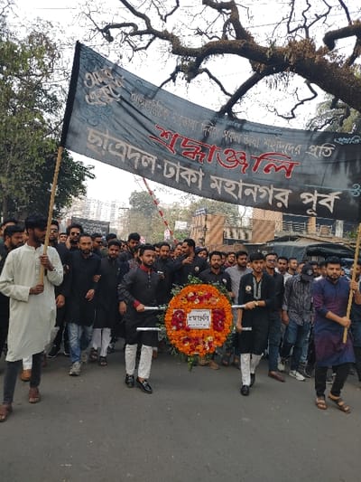 A group of people in Dhaka, Bangladesh, are marching with a banner written in Bengali. The banner appears to be for the student wing of a political party, "Chhatra Dal", and commemorates an event or leader. They are carrying a large, circular floral wreath with a central inscription in Bengali. The march is taking place outdoors on a street during daytime. Trees are visible on the sides, suggesting an urban setting.
