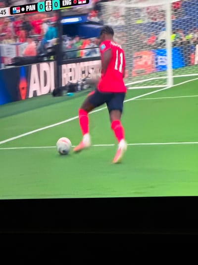 The image shows a cropped view of a soccer match, likely from a television broadcast. A player in a red jersey with the number 11 on the back is actively playing the ball. He is wearing dark blue shorts and bright pink socks. The player is on a green soccer field with white lines. A soccer ball is visible near his feet. In the background, there are blurred spectators and a goal net. A scoreboard at the top of the image shows "PAN 0 0 ENG", indicating the teams playing and the current score. The location is not specified as Moscow, Russia in the provided image or crops.