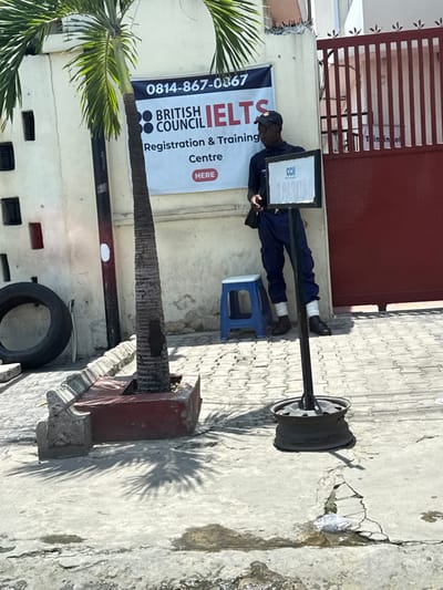 The image depicts an outdoor scene at the entrance of a facility identified by a banner as a "BRITISH COUNCIL IELTS Registration & Training Centre" in Alaguntan, Nigeria.
In the foreground, a concrete surface shows cracks and small puddles. A slender palm tree grows from a rectangular red concrete planter box on the left. To its left, a black vehicle tire rests against a light-colored building wall. To the right of the palm tree, a small, blue plastic stool is positioned on a paved area composed of interlocking blocks.
A black-framed banner is affixed to the building wall, displaying the phone number "0814-867-0367" and the text "BRITISH COUNCIL IELTS Registration & Training Centre," with a red box containing "HERE."
An adult male stands leaning against the wall, to the right of the banner and to the left of a large red metal gate that occupies the right side of the frame. He has dark skin, wears a dark baseball cap, a dark blue short-sleeved shirt, dark blue cargo pants, and dark shoes with white material visible above the ankles. A dark bag is slung over his left shoulder. He holds a small, dark object, possibly a mobile phone, in his left hand and gazes towards the left of the frame.
In the middle foreground, a dark metal pole with a rectangular sign attached stands anchored by a metallic wheel rim. The sign displays "CCII" at the top, with additional obscured text below. Shadows from the palm tree and the sign stand are cast on the ground, indicating bright overhead sunlight. A piece of white litter is visible on the cracked concrete near the sign's base.