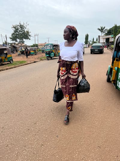 In Bukuru, Nigeria, a woman walks on a paved road, carrying two black bags. She is wearing a white shirt and a patterned maroon skirt, with a matching headscarf. The scene is bustling with activity, featuring several auto-rickshaws, also known as Keke Napeps, parked and in motion. A green sedan is visible on the road behind her, and a building with the name "KUGEL HOTEL RESORT" is in the background. Power lines crisscross the sky above the various structures and vegetation.