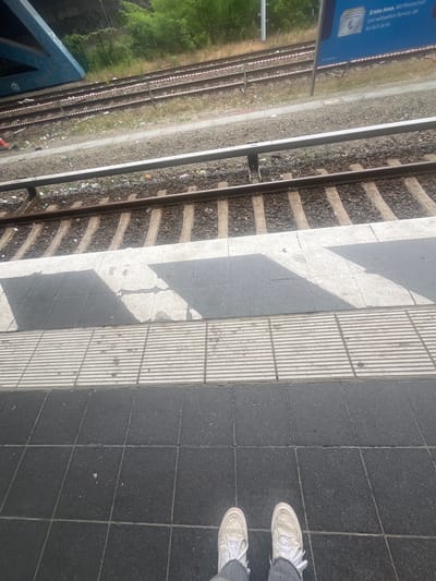 This image depicts a ground-level view of a train station platform in Berlin, Germany, looking down at the train tracks and the edge of the platform. In the foreground, a person's feet clad in white sneakers and gray pants are visible, standing on a dark tiled surface. The platform edge is marked by a textured, raised strip, followed by a section of dark and light patterned tiles. Beyond the platform, train tracks run parallel, with gravel ballast and concrete sleepers. Some debris, including plastic wrappers and cigarette butts, is scattered along the tracks and platform edge. In the background, a blue train carriage is partially visible, along with overgrown green vegetation and a section of a blue overhead structure with graffiti. A blue advertisement poster with German text is also visible on a pole to the right of the tracks. The overall scene suggests an urban outdoor environment at a public transportation hub.