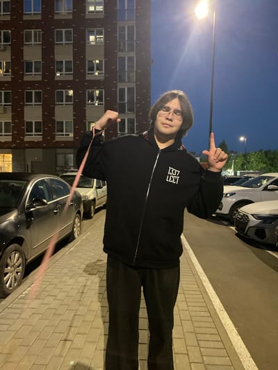 A young man with dark hair and glasses stands on a paved sidewalk in front of a brick apartment building at dusk. He is wearing a black zip-up hoodie with a white logo that reads "1337 LEET" on the chest, and dark pants. His right arm is raised, with his index finger pointing upwards. A pink strap hangs from his left hand, extending towards the ground. Parked cars are visible behind him on the right, and a darker car is parked on his left. A lamppost illuminates the scene. The location is the courtyard of an apartment complex in Izhevsk, Russia.