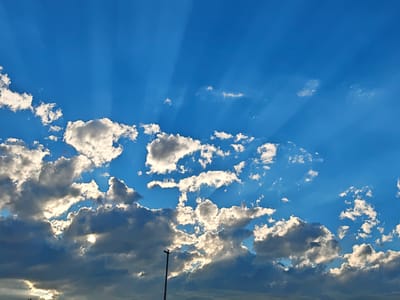 The image shows a vibrant blue sky with scattered clouds. Rays of sunlight are visible, piercing through the clouds, creating dramatic lighting effects. The clouds vary in density, with some appearing bright white and others darker grey, suggesting different levels of light penetration. In the lower part of the frame, a silhouette of a lamppost is visible against the bright sky. The scene is a landscape view of the sky, likely taken from an outdoor vantage point. There are no people or specific landmarks clearly identifiable within the provided image to pinpoint a precise location within Tbilisi, Georgia.