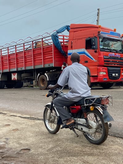 The image captures a street scene in Mararaba, Nigeria, featuring a man on a motorcycle in the foreground and a large red truck in the background.

In the foreground, a bald Black male, viewed from behind, is seated on a red and chrome motorcycle. He is wearing a light grey short-sleeved collared shirt and dark grey trousers, with dark sandals visible on his feet. The motorcycle features chrome handlebars, spoked wheels with a white front fender, a black seat, and a chrome rear rack equipped with four circular red taillights/reflectors and a single yellow circular turn signal on the right side. A kickstand is visible on the left side, and an exhaust pipe is visible on the right.

In the midground and background, a large red DAF XF tractor-trailer is prominently displayed. The truck's cab is red with blue and white stripe decals along its side and features "ماشا الله" (Masha'Allah) written in Arabic script above the windshield. Amber warning lights are mounted on the cab's roof. The trailer is a red flatbed with high metal sides, reinforced with an upper rail structure. A blue tarp is partially draped over the front upper left side of the trailer. Inside the trailer, light-colored material, possibly hay or sacks, is visible near the front. A white and red 'X' hazard marking is present on the lower rear side of the trailer, and a spare tire is mounted underneath. Two other individuals are present near the truck: one person with dark skin, wearing a white shirt with red trim, is partially visible standing inside the trailer bed near the cab, and another individual, also with dark skin and wearing a dark shirt, is partially visible near the truck's passenger side door.

The road surface is a greyish-brown paved material, exhibiting numerous cracks and minor depressions. The sky above is overcast, presenting a uniform grey tone. A wooden utility pole with multiple overhead wires is visible behind the truck cab.
