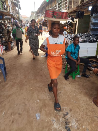 In a bustling marketplace located in Fegge, Nigeria, a young person wearing an orange smock with the words "Mega Supermarket" walks down a dirt path with items balanced on their head. A woman in a patterned dress is walking in the same direction, and a person with a green backpack is walking toward the camera. To the right, a person in a blue shirt sits on a green stool near a stall of shoes. Other people and stalls are visible in the background, illustrating a busy market scene.