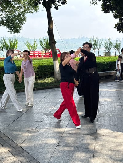 In Jiangjin, China, several women are engaged in a dance session outdoors on a paved walkway. A woman in a blue top and white pants is on the left, holding hands with a woman in a purple top and white pants. Further to the right, two other women are dancing together. One, in a black top and red pants, is in the foreground. Another woman in a black outfit with a belt is to her right. In the background, a red vehicle is parked, and lush green bushes and trees line the area. The sky is overcast, and the ground is a patterned grey stone pavement.