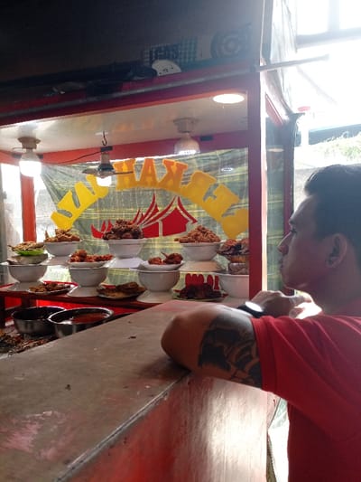 The image depicts a food stall, likely a "ayam penyet" stall, located at the coordinates -6.987, 107.553 in Katapang, Indonesia. The stall is made of red metal and features a yellow sign with the words "Ayam Penyet" written in yellow letters. It is partially enclosed with glass windows, with a brown wooden countertop. The stall is stacked with platters of food, likely containing deep-fried chicken and other accompanying dishes. A single man, wearing a red t-shirt, a black watch, and a tattoo on his left arm, is leaning on the countertop with his back to the camera, gazing at the food. His expression is contemplative. Behind him, a large window reveals a partially obstructed view of the street outside. The scene is lit by artificial lighting, casting a warm glow on the stall and the food.