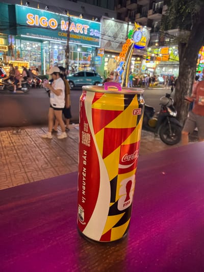 This media file is a close-up, sideways shot of a Coca-Cola can with a distinct, colorful design, positioned in the foreground. The can features a pattern of red, yellow, and black geometric shapes, along with the Coca-Cola logo and "FIFA" text, suggesting a promotional collaboration. Part of a purple surface occupies the right side of the frame, possibly a table.
In the background, a busy street scene unfolds at night. Neon lights illuminate storefronts and signs, creating a vibrant, urban atmosphere. A prominent sign reads "SIGO MART 24H CONVENIENT EVERYDAY," indicating a convenience store. Several motorcycles are parked along the street, and people can be seen walking and interacting in front of the store. The presence of motorcycles and the signage in English and what appears to be Vietnamese suggest a location in Vietnam. The overall impression is a lively evening in a commercial area, likely a city street. The lighting suggests it is nighttime, with artificial lights dominating the scene. No specific weather conditions are evident.