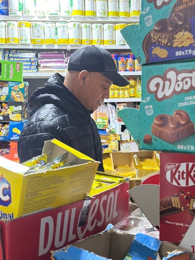The scene depicts the interior of a retail store, identified as being in the city of Meknes, Morocco. An adult male is visible in profile, facing towards the right side of the frame. He wears a black quilted puffer jacket and a black mesh-back baseball cap. His head is tilted downwards, suggesting he is examining merchandise.
The environment features multiple display shelves and stacked product boxes. In the immediate foreground, a red cardboard display box for "Nestlé Kit Kat" is partially visible on the right, alongside a larger red box with "DULCESOL" printed on it. A yellow, partially open cardboard box containing smaller yellow packages rests above the "DULCESOL" box. To the right of the male subject, two stacked turquoise-colored display boxes for "Wooly" brand milk chocolate are prominent, with text indicating "CORN FLAKES" and "MILK CHOCOLATE" along with "80x7,5g" packaging details.
In the background, shelves are fully stocked with various packaged goods. Rows of cylindrical products, likely rolls of paper, with white and yellow packaging are visible on upper shelves. Lower shelves contain items such as bags of chips or snacks, bottles (some yellow, potentially cooking oil), and other general grocery items. Below the "Wooly" displays, open bins hold a loose yellow granular product. The store appears to be well-lit by overhead lighting.