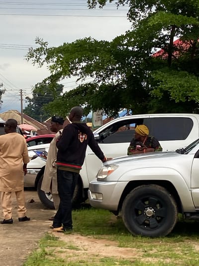The image depicts an outdoor street scene in Mararaba, Nigeria, under an overcast sky. Four individuals are present near parked vehicles on a dirt ground with sporadic grass patches. From left to right, a male in a tan traditional outfit stands with his back to the viewer. Adjacent, a second male in a light-colored traditional top and black cap is shown in right profile. A third male, wearing a black hoodie featuring a large lion emblem on the back, has his right arm resting on the hood of a silver SUV. He faces a female in a patterned top and yellow headwrap, who is leaning out of the passenger side window of an adjacent white SUV, looking towards him. Utility poles and power lines are visible against the sky. The background consists of dense green tree foliage partially obscuring simple structures with corrugated metal roofs, including one prominent red roof.