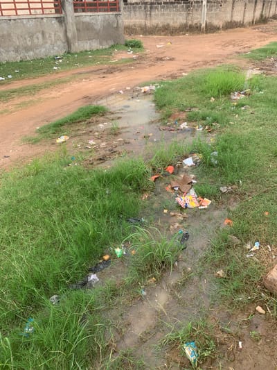In Lafia, Nigeria, a muddy area with standing water is visible. The ground is uneven and appears to be a dirt path or roadside, littered with a significant amount of trash. Among the debris are plastic wrappers, paper packaging, and discarded food items. Lush green grass grows around the edges of the muddy patches. In the background, a rough concrete wall with a red-barred structure runs along the top. Further back, a brick building with a rough texture is visible. The scene suggests an area with poor waste management and potential drainage issues.