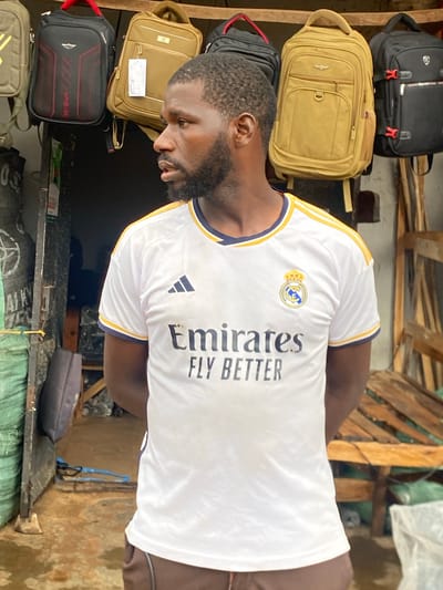 A man stands in a market stall in Bukuru, Nigeria, wearing a white Real Madrid jersey. He has dark skin, a beard, and short hair, and he is looking to his right. Behind him, several backpacks are hanging on display, in various colors including black, tan, and gold. The stall appears to be open-air, with a dirt floor and wooden shelves. The background shows a dark doorway and some wooden structures.