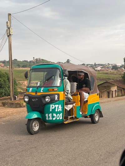 A man is lying down inside a green and yellow auto-rickshaw, appearing to be asleep or resting. He is wearing a black t-shirt and white shorts. The auto-rickshaw is parked on a paved road, with a rural landscape featuring hills, scattered buildings, and vegetation in the background. A utility pole with several electric meters is visible on the left side of the image. The sky is overcast, suggesting it might be morning or late afternoon on a cloudy day. The text "PTA 11301" is visible on the side of the auto-rickshaw, along with the word "CHEER" above the headlights. The overall scene depicts a moment of rest for the driver of the auto-rickshaw in a roadside setting in Jos, Nigeria.