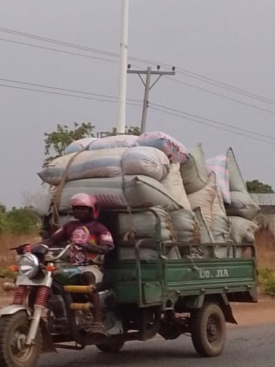The image depicts a three-wheeled motorcycle, heavily laden with sacks, being driven by a person on a road in Dungu, Ghana. The driver, an adult male, is wearing a pink helmet and a dark t-shirt featuring a graphic of a face.

The vehicle is a motorcycle at the front, attached to a large, deep green cargo bed at the rear. This cargo bed is piled high with numerous large sacks or bags, predominantly light-colored (white, cream, light blue), some with visible pink and blue stripes. The load is substantial, towering above the driver, and appears to be secured with ropes.

The scene is set outdoors under an overcast, grey sky, indicating daytime with no direct sunlight. In the background, a tall white pole and power lines stretch across the sky, with distant green foliage visible on the horizon. The road surface beneath the vehicle appears to be unpaved or a dirt track.

Visible text on the side of the green cargo bed reads "UO. JIA". The image captures a common method of transporting goods in the region, emphasizing the heavy load capacity of such vehicles.