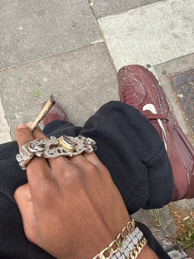 A person wearing a brown Nike Air Max 90 sneaker, black sweatpants, and several bracelets and a large ring is sitting on a sidewalk smoking a hand-rolled cigarette. The sidewalk appears to be made of concrete slabs. The location is in London, United Kingdom.