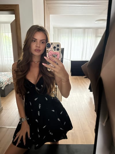 A young woman with long, brown, wavy hair is taking a mirror selfie in a room. She is wearing a black mini dress with a floral pattern and a thin silver necklace. She has a watch on her left wrist and her fingernails are painted light pink. She is holding a phone with a decorative case in her right hand, which is visible in the mirror. The room has wooden floors, white sheer curtains on the windows, and a bed with a patterned comforter in the background. The reflection in the mirror also shows a doorway and a closet. The scene appears to be an indoor residential setting, possibly an apartment or house. There is no information in the image to suggest a specific location in Plovdiv, Bulgaria.