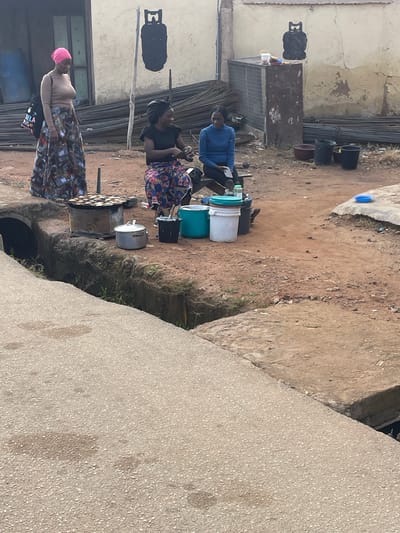 In Rafin Par, Nigeria, an outdoor scene unfolds with three women engaged in activities related to food preparation and vending. A woman on the left, wearing a pink headscarf and a long, patterned skirt, stands near a cooking stove laden with small, round food items. Beside her, a black bag with colorful lettering is slung over her shoulder. Two other women are seated. One, dressed in a black top and a floral skirt, appears to be attending to something in front of her. The other, in a blue long-sleeved shirt and dark pants, is seated and holds what appears to be a mobile phone. The environment is characterized by a dirt ground, with stacks of metal rods and construction materials visible in the background. A concrete structure, possibly a building, forms the backdrop. Several buckets and pots are placed around the women, suggesting their use in the food vending operation. A drainage channel runs along the foreground, with a concrete walkway alongside it. The overall impression is a snapshot of daily commerce in a Nigerian urban setting.