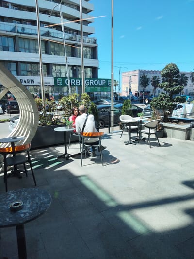 The image depicts an outdoor cafe scene in Batumi, Georgia, on a sunny day. Two people, a man and a woman, are seated at a small round table, engaged in conversation. The man is wearing a white shirt with an orange strap over his shoulder, while the woman has on a red jacket. Behind them, a planter box is filled with greenery. To the left is a part of a circular architectural structure. Other tables and chairs are scattered around the patio area. Behind the cafe is a modern, multi-story building with signage for "PARIS," a shop, and "ORBI GROUP" visible. Across the street, another building, in the style of a castle, can be seen. Several parked cars are visible on the street. The ground is paved with grey stone tiles. The image is taken at ground level.