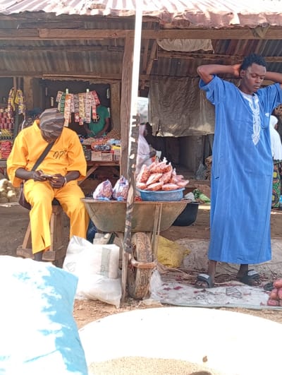 This media file captures a scene at an outdoor market in Wamba, Nigeria. Two men are prominently featured. The man on the left is seated on a wooden stool, wearing a bright orange traditional outfit and a striped headscarf. He is engrossed in his mobile phone, with a dark strap from a bag slung across his shoulder. The man on the right stands, wearing a long blue traditional robe with white embroidery and dark sandals. He has his hands clasped behind his head, seemingly in a relaxed or pensive pose.
The setting is a market stall with a rustic, corrugated metal roof supported by wooden beams. Behind the seated man, various goods are displayed on shelves, including packaged snacks and what appear to be food items in cans and plastic bags. A green shirt is visible on another person in the background, partially obscured. In the foreground, a wheelbarrow holds a blue basket filled with packaged goods, possibly food items. Large sacks, one white and one yellow, are placed near the wheelbarrow. The ground is dirt and scattered with some red debris. The lighting suggests it is daytime, likely with strong sunlight given the shadows. The overall atmosphere appears to be that of a busy, functional marketplace.