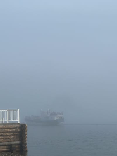 A ferry is traveling on a foggy body of water. The ferry has multiple decks and people are visible on the top deck. In the foreground, there is a wooden dock with a white railing. The water is calm and gray, and the fog creates a hazy atmosphere. The scene appears to be in Listvyanka, Russia.
