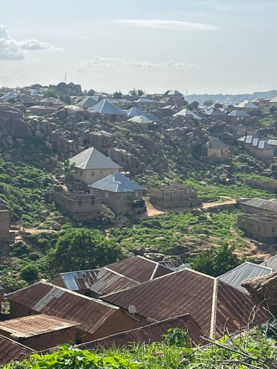 The image depicts a view of a residential area nestled on a rocky, hilly terrain. The architecture consists primarily of low-rise buildings with corrugated metal roofs, many of which appear weathered and rusted. Some structures are under construction, with visible brickwork and incomplete walls. Green vegetation, including grass and trees, is scattered across the landscape, growing amongst the rocks and around the buildings. The sky is overcast with diffused sunlight. The overall impression is of a community built into a challenging, natural environment. The style of construction and the landscape are consistent with the rocky hills and settlements found in and around Jos, Nigeria.