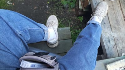 The image shows a first-person perspective of someone sitting outdoors on wooden planks, likely a bench or deck. The person is wearing blue jeans and white socks with light-colored sneakers. A small, light-colored bag with a zipper is visible on the lap, with a white box, possibly a cigarette pack, protruding from it. The environment appears to be rural or suburban, with grass and dirt visible in the background, and some stacked wood or debris. The lighting suggests it is daytime. The location is identified as Yudin, Russia.