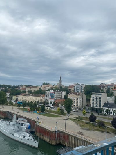 The image captures a cloudy day overlooking the Sava River in Belgrade, Serbia. In the foreground, the bow of a gray ship labeled "SAVA" is docked. Behind the ship, a wide promenade area with paved surfaces and green spaces is visible. A playground with colorful equipment is situated on the promenade.

Beyond the promenade, a city skyline rises, characterized by a mix of architectural styles. Numerous buildings, varying in height and color, are nestled amongst lush green trees. Some buildings exhibit classical European architecture with ornate facades, while others are modern high-rises. A prominent church steeple with a green dome can be seen in the distance, suggesting a religious or historical landmark. Various vehicles, including buses and cars, are visible on the road running parallel to the promenade. The sky is overcast with heavy gray clouds, creating a subdued and diffused light. The overall scene depicts a riverfront area in a European city, likely a historic district due to the blend of old and new architecture.