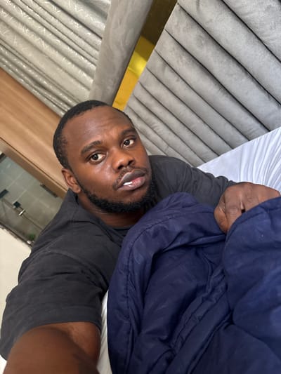 A man with dark skin, a beard, and a black t-shirt takes a selfie indoors. He is partially covered by a dark blue puffy jacket. The background includes a grey, upholstered headboard with gold trim, a white sheet, and a glimpse of a bright, possibly tiled, room. The location, Iddo Tudunwada, Nigeria, cannot be confirmed from visual information.