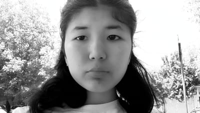 This is a black and white, close-up, front-facing portrait of a young East Asian female taken outdoors in Osh City, Kyrgyzstan. She is wearing a white t-shirt and small, sparkling stud earrings. Her hair is dark and pulled back, with some strands falling around her face. The background consists of trees with visible leaves and a bright, overcast sky. A wooden pole with a sign is visible to the right. The lighting suggests it is daytime.