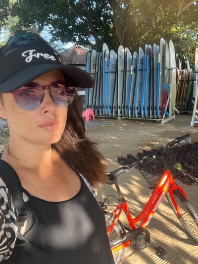 The image shows a woman in a black visor, sunglasses, and a black tank top, taking a selfie. Behind her, a rack of surfboards stands on a sandy beach. A red bicycle with a basket is parked in the foreground. In the background, there is a small pink stool and trees. The scene is lit by sunlight, casting shadows on the ground. The presence of surfboards and the sandy beach suggests a coastal location, likely in Kuta Selatan, Indonesia, a popular tourist destination known for its beaches and surf spots.