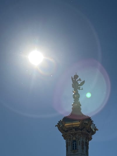 A close-up, low-angle shot captures the sun, appearing as a bright, white orb with rays extending outward, in a clear, blue sky. Lens flare creates colorful rings and streaks of light across the image. In the lower right portion of the frame, the top of a building's ornate spire is visible. The spire is decorated with intricate gold detailing and a red roofline. At the very top of the spire stands a silhouette of a two-headed eagle, a common symbol in Russian heraldry, wearing a crown. The location is identified as Peterhof, Russia, likely referring to a prominent architectural feature of the Peterhof Palace. There are no people or active interactions depicted in the image, only the static architectural element against the bright sky.