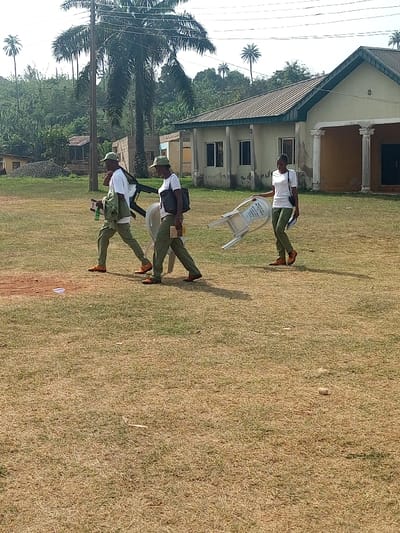 This image captures a daytime scene in Aramoko-Ekiti, Nigeria, featuring three individuals walking across a dry, patchy grass field from left to right. All three are uniformly dressed in white t-shirts, olive-green trousers, and brown shoes; the leading male and the female behind him also wear olive-green caps. The male on the left carries a white foldable plastic chair on his left shoulder, a green bottle in his left hand, and an olive-green jacket draped over his arm. The female immediately to his right carries a dark shoulder bag on her left shoulder, a booklet or papers in her right hand, and a white foldable plastic chair partially visible by her left side. The third individual, a female on the far right, holds a white foldable plastic chair horizontally in her right hand and papers in her left. In the background, extensive green foliage and palm trees rise, accompanied by a utility pole. A light-colored, single-story building with a gabled roof and two prominent white columns is situated in the mid-right background, adjacent to partially constructed or weathered structures.