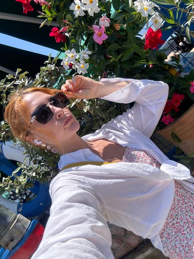 The image shows a woman with red hair wearing sunglasses, a white unbuttoned shirt, and a floral-patterned dress. She is holding her sunglasses with her left hand and has white wireless earbuds in her ears. She is leaning against a railing that has a blue and red structure attached to it, and a plant with red, white, and pink flowers is growing around her. The background appears to be outdoors, with sunlight casting shadows. The setting resembles a public space with greenery and some industrial-looking elements. Given the presence of a yacht club structure in the background and the overall aesthetic, this scene is likely captured at the Budva Yacht Club in Budva, Montenegro, a popular coastal city known for its scenic beauty and vibrant atmosphere.
