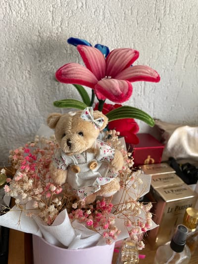 A close-up shot features a small, light brown teddy bear wearing a floral-patterned jumpsuit and a matching bow in its ear. The bear is nestled among a bouquet of dried baby's breath flowers, some with pink accents, which are presented in a pink container lined with white paper. In the background, a large, artificial pink and red flower with blue accents stands tall. To the right, a red gift box is visible, along with several gold-colored perfume boxes labeled "Le Parfum." A glass spray bottle and other perfume bottles are also present on the surface. The scene is illuminated by natural light, casting soft shadows. The image is likely from Zlatograd, Bulgaria, judging by the context provided.
