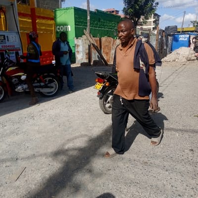 A man with dark skin and wearing a brown polo shirt, dark pants, and sandals walks on a cracked asphalt road. He has a navy blue jacket draped over his shoulder. Behind him is a black motorcycle. Further back, two other men stand near a gas pump and a green building with white letters. A "FRESH MILK" sign is visible in the background. The sky is partly cloudy.