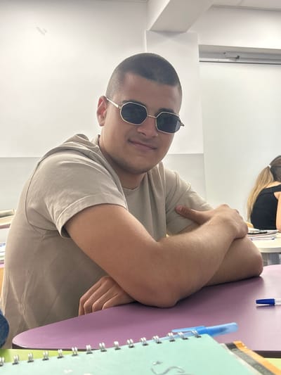 The image shows a young man wearing sunglasses and a tan t-shirt, sitting at a desk. His arms are crossed in front of him. The desk is covered with notebooks and pens. The background appears to be a classroom or lecture hall, with whiteboards and other students visible. The location is identified as Sliven, Bulgaria.